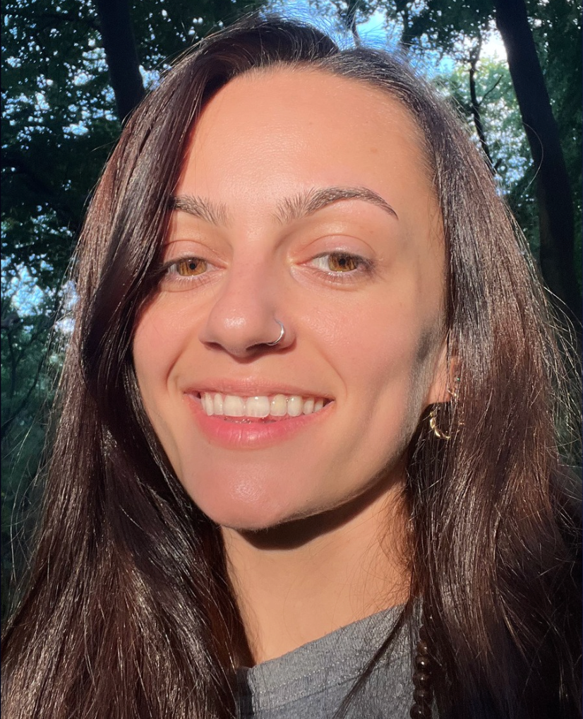 Woman with long brown hair smiling outdoors with trees in the background